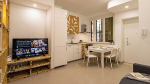 Una cocina moderna con armarios blancos, una mesa de comedor con sillas y un televisor mostrando 'GINNY & GEORGE'.