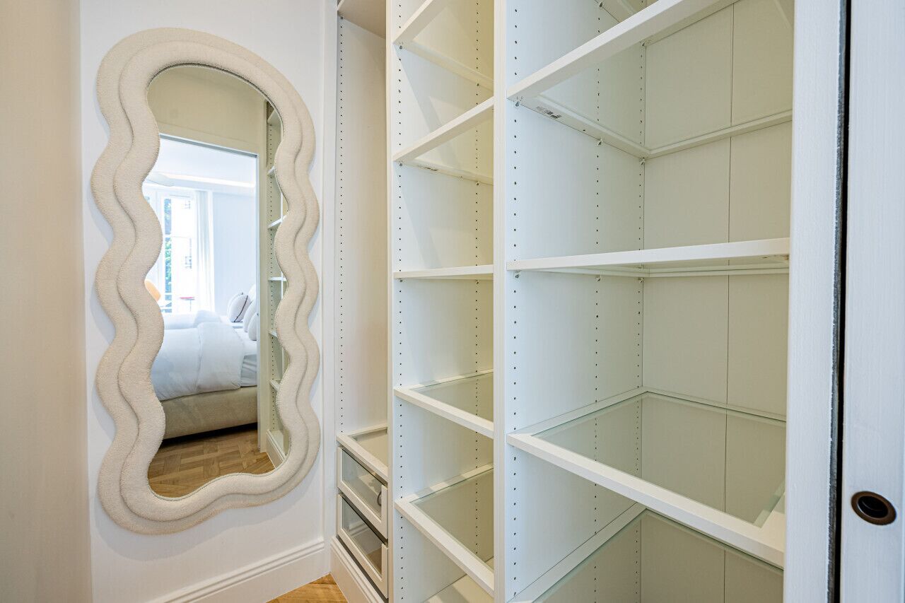 A room with a large mirror, a bed, and a white wardrobe with multiple shelves.