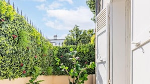 Uma varanda com cadeira de vime e uma mesa pequena, rodeada por vegetação exuberante e um edifício branco.