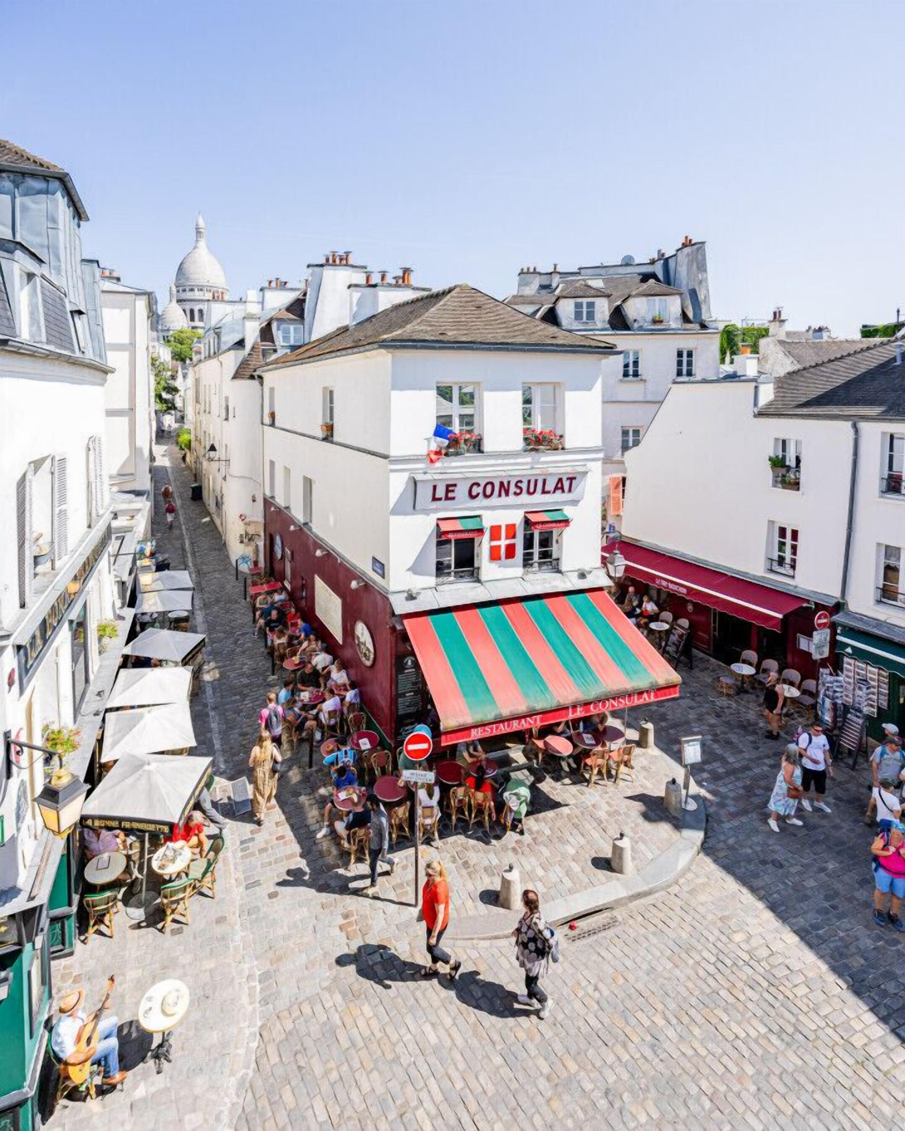 En livlig gatuscen med en framträdande restaurang som heter ”LE CONSULAT” och som har ett randigt markis.