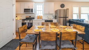Una cocina moderna con armarios blancos, un refrigerador de acero inoxidable y un comedor preparado para seis personas, con sillas de madera y cojines grises.