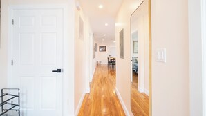 Un pasillo con piso de madera, paredes blancas y una puerta de color blanco. Un espejo se encuentra en la pared derecha y un zapatero en la izquierda.