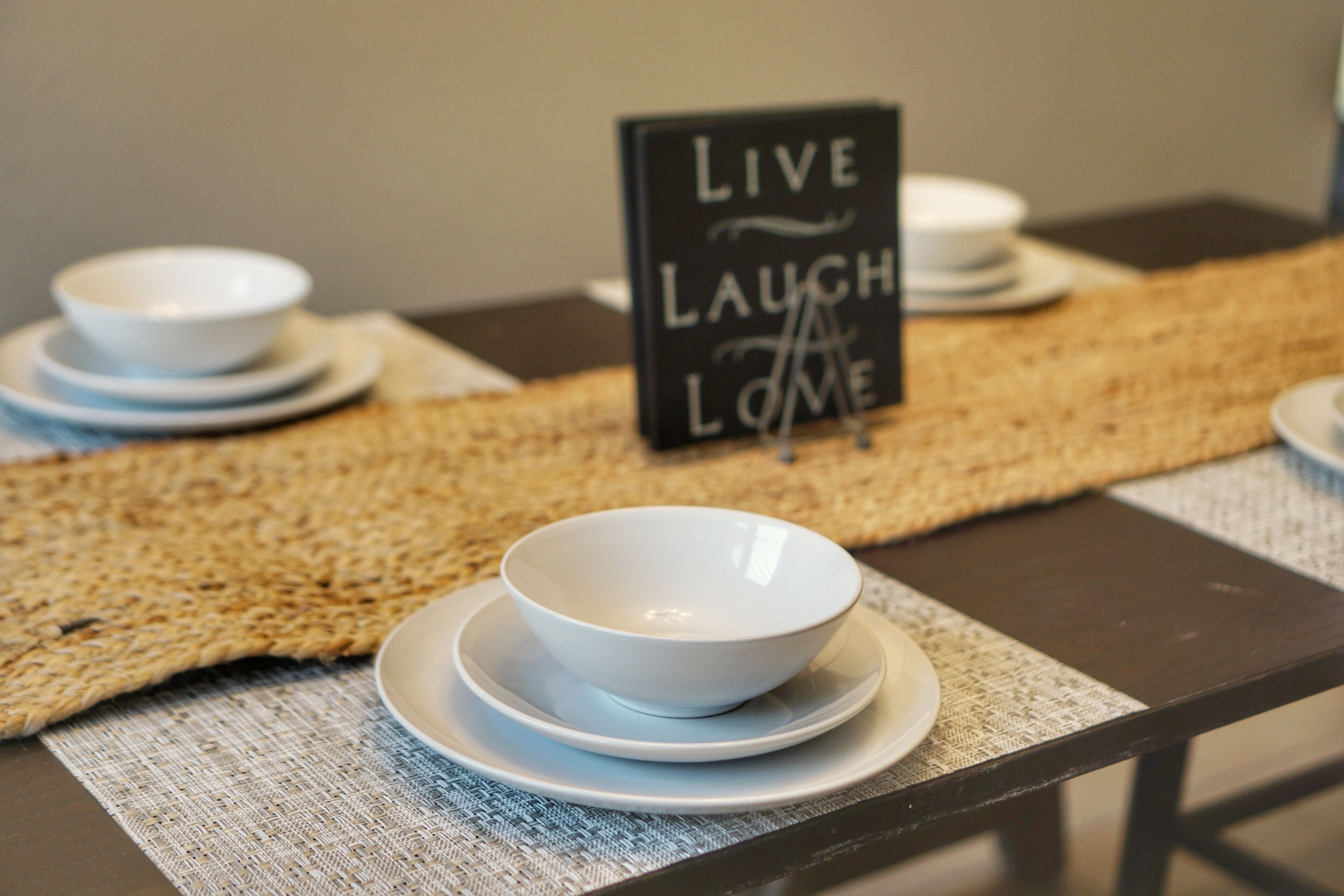 A table with white bowls and plates, a woven placemat, and a sign that says "LIVE LAUGH LOVE".