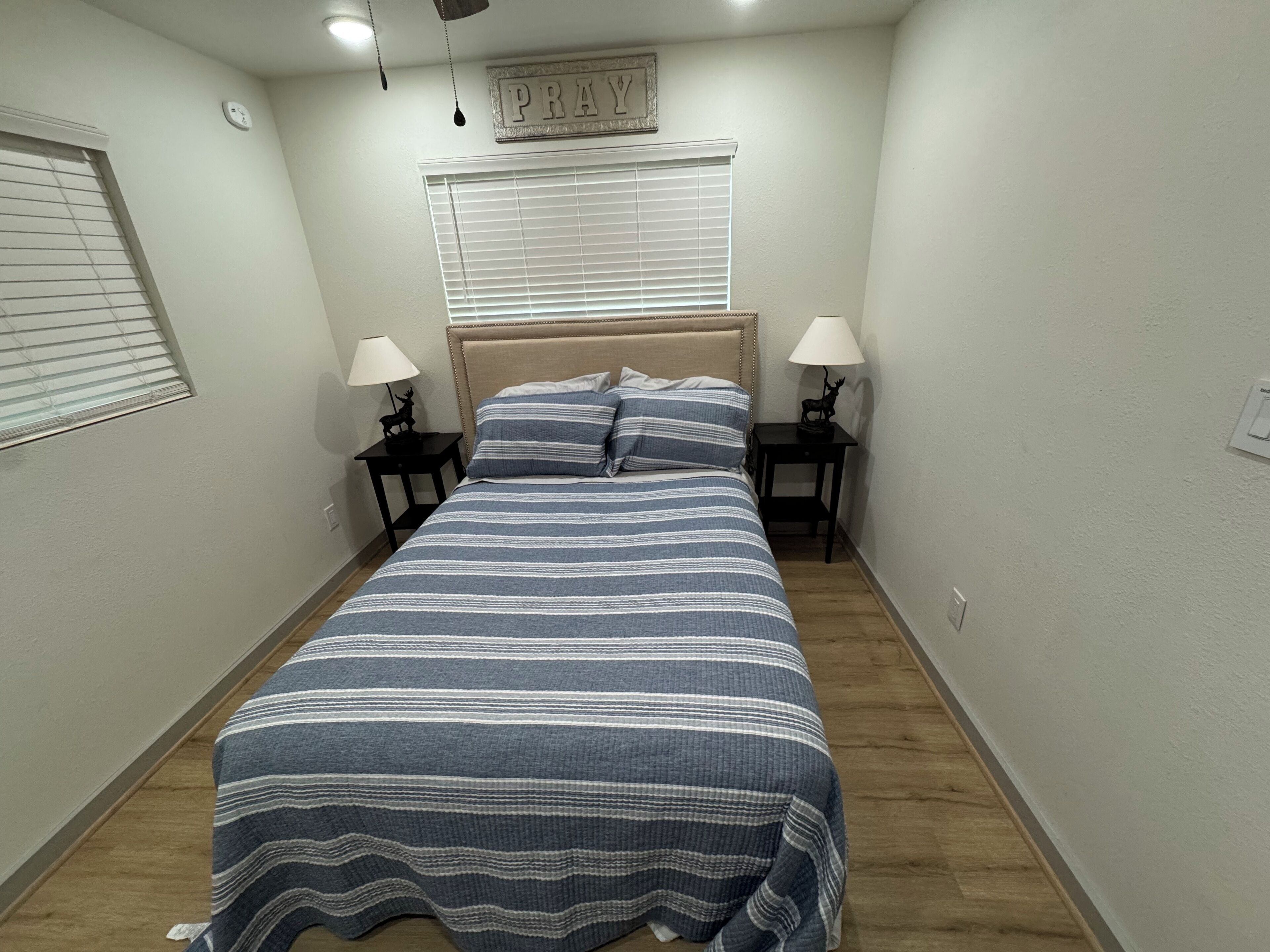 A bedroom with a bed, two bedside tables, a window with blinds, a wall clock, and a 'PRAY' sign above the bed.