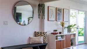 Un salon moderne avec un grand miroir rond, un banc, un meuble blanc rempli de livres et, au mur, des œuvres d’art abstraites encadrées.