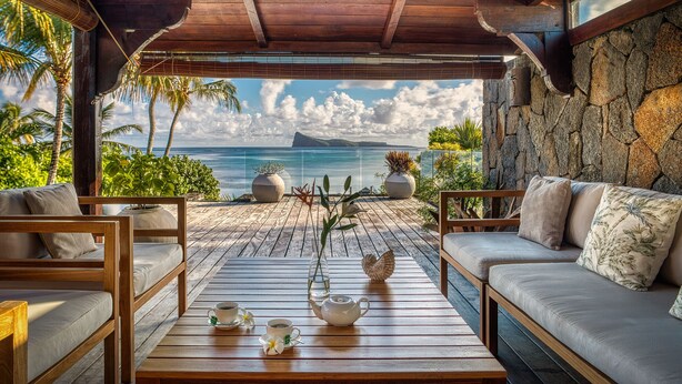 A wooden deck with a stone wall, a cushioned sofa, and a table with a teapot and cups. - Villa Zébra Moon by muse villas  (Bain Boeuf)