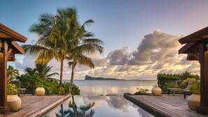 A wooden deck with a pool reflecting palm trees and a sunset over the ocean. - Villa Zébra Moon by muse villas  (Bain Boeuf)