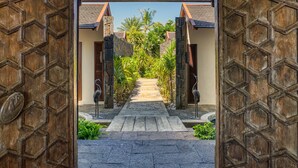 A wooden entrance with a stone pathway leading to a building with a tiled roof and a view of greenery. - Villa Zébra Moon by muse villas  (Bain Boeuf)