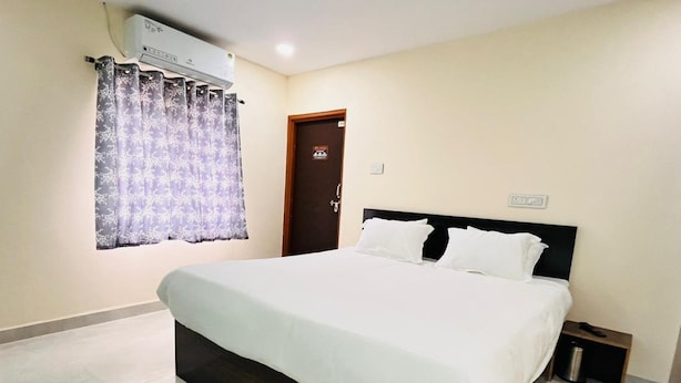 A hotel room with a large bed, a small bedside table, a nightstand, a window with a curtain, and an air conditioning unit above the window. - Hotel Grand Imperial (Chhatrapati Sambhajinagar)