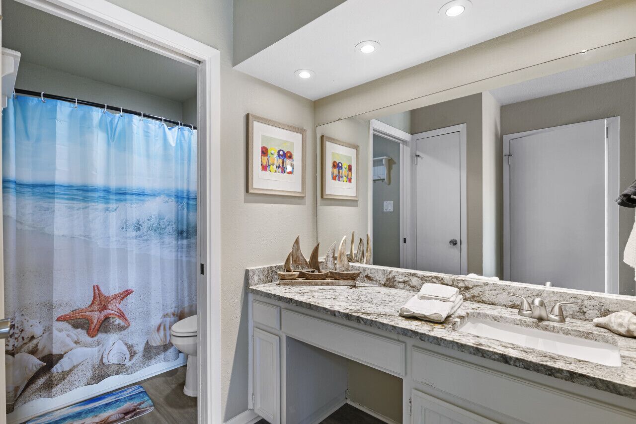 A bathroom with a shower curtain depicting a beach scene, a marble sink, and framed artwork on the wall.