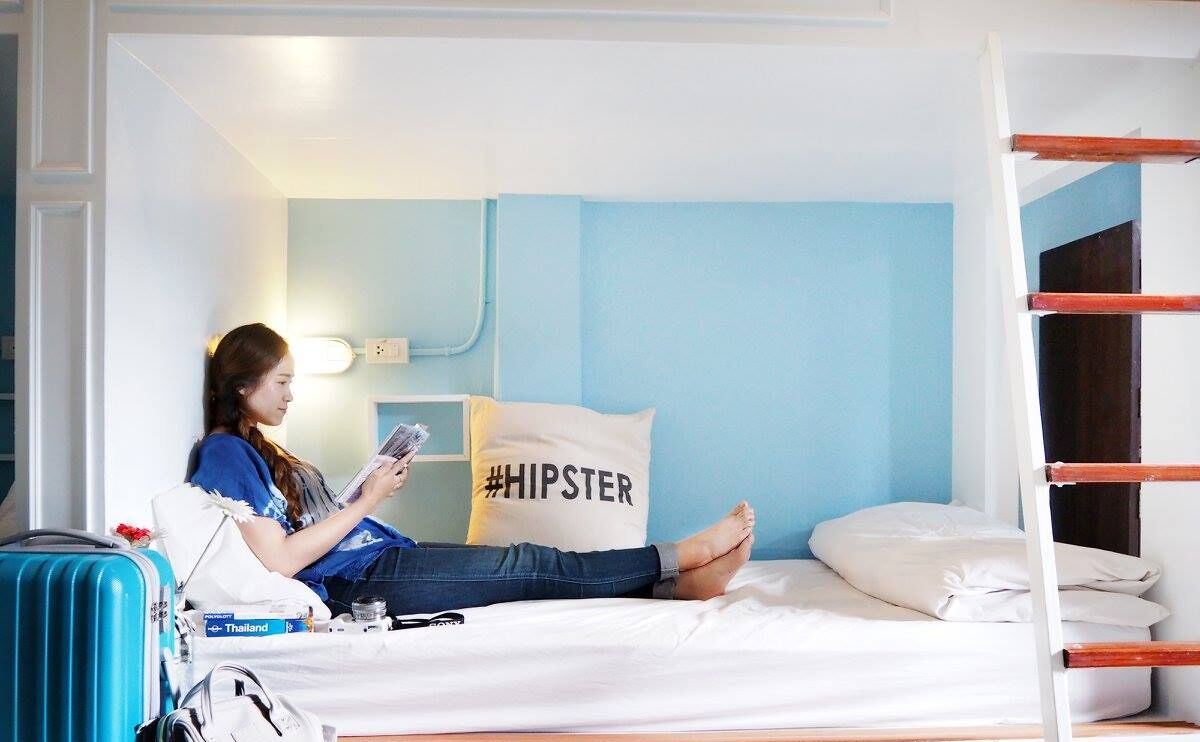 A room with a bunk bed, a suitcase, a pillow with #HIPSTER, and a wall-mounted light.