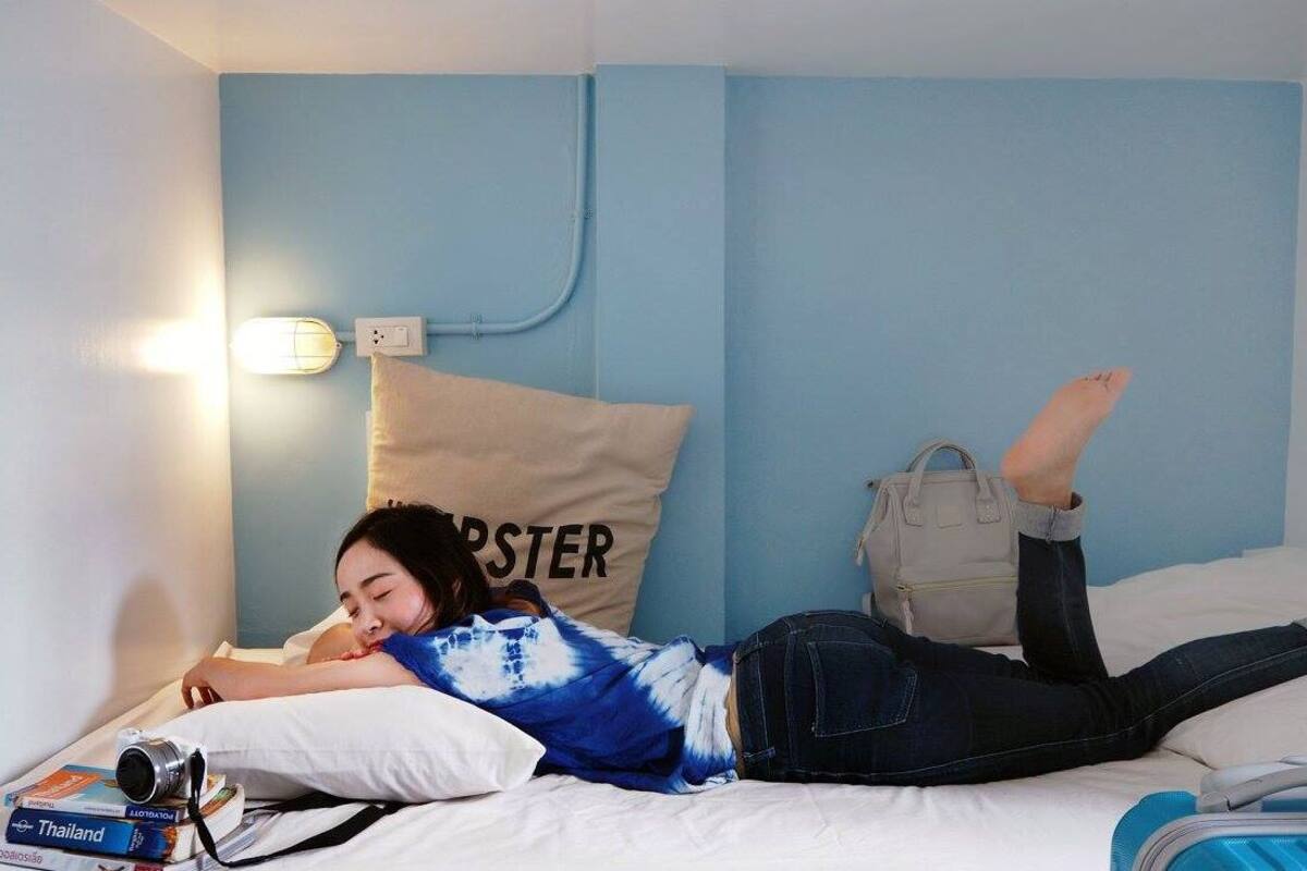 A person lying on a bed in a room with a blue wall, a white pillow with the word "ROSTER" on it, a white lamp, and a bag on the bed.