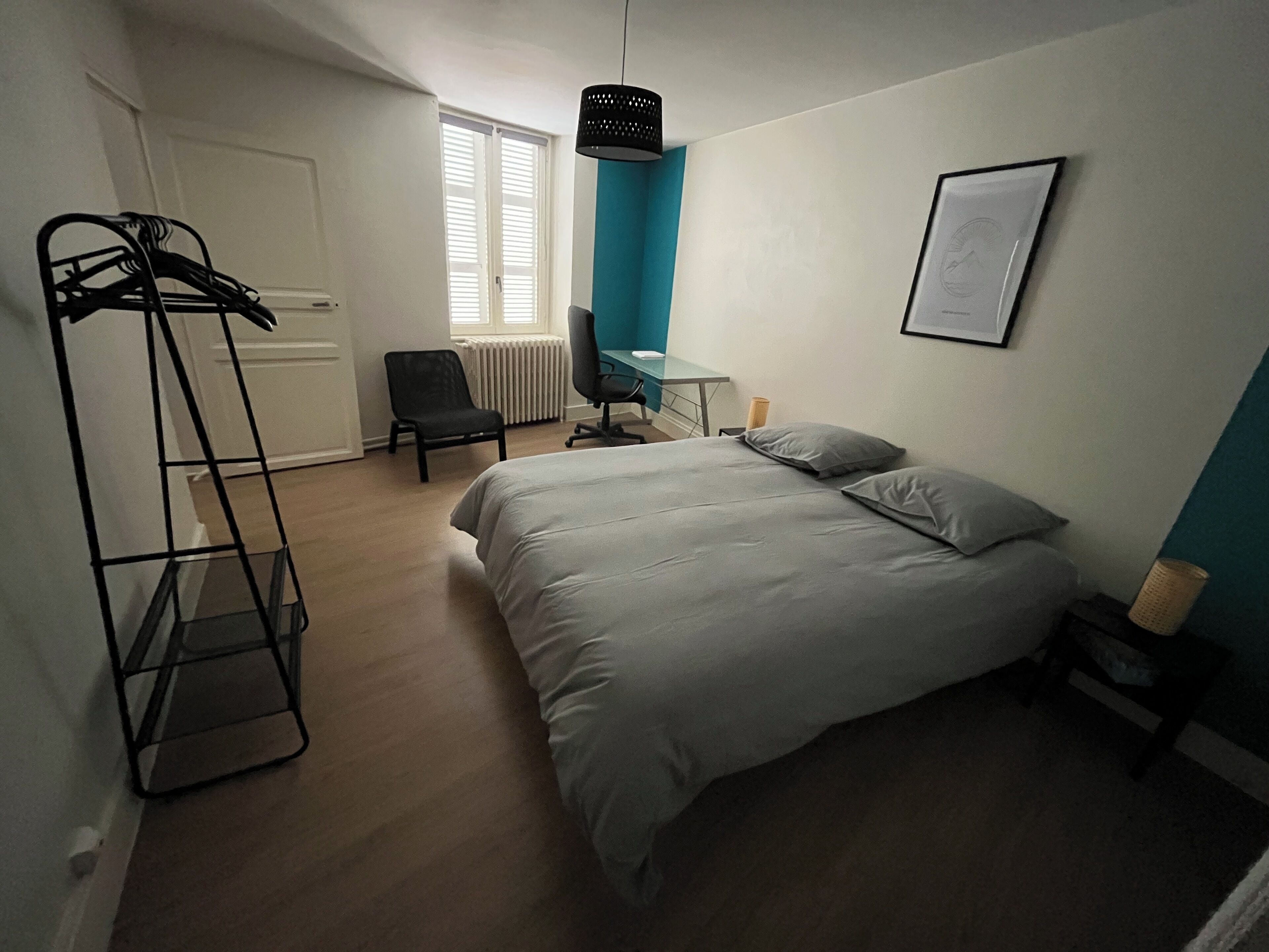 A bedroom with a bed, a chair, a desk, a radiator, and a hanging light fixture.