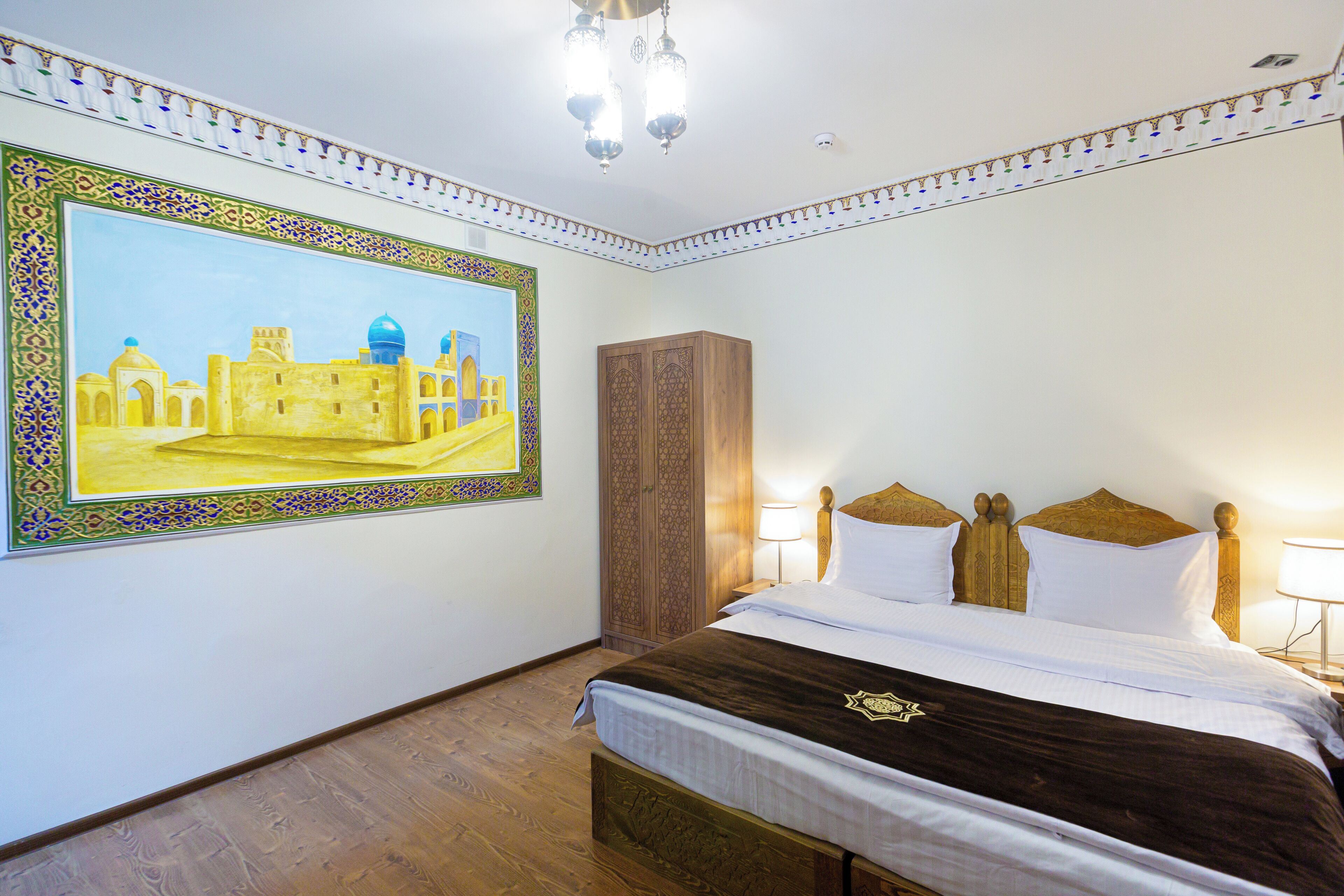 Une chambre d’hôtel avec un grand tableau représentant un bâtiment historique, une armoire en bois, un lit avec une literie blanche et marron, et un lustre.