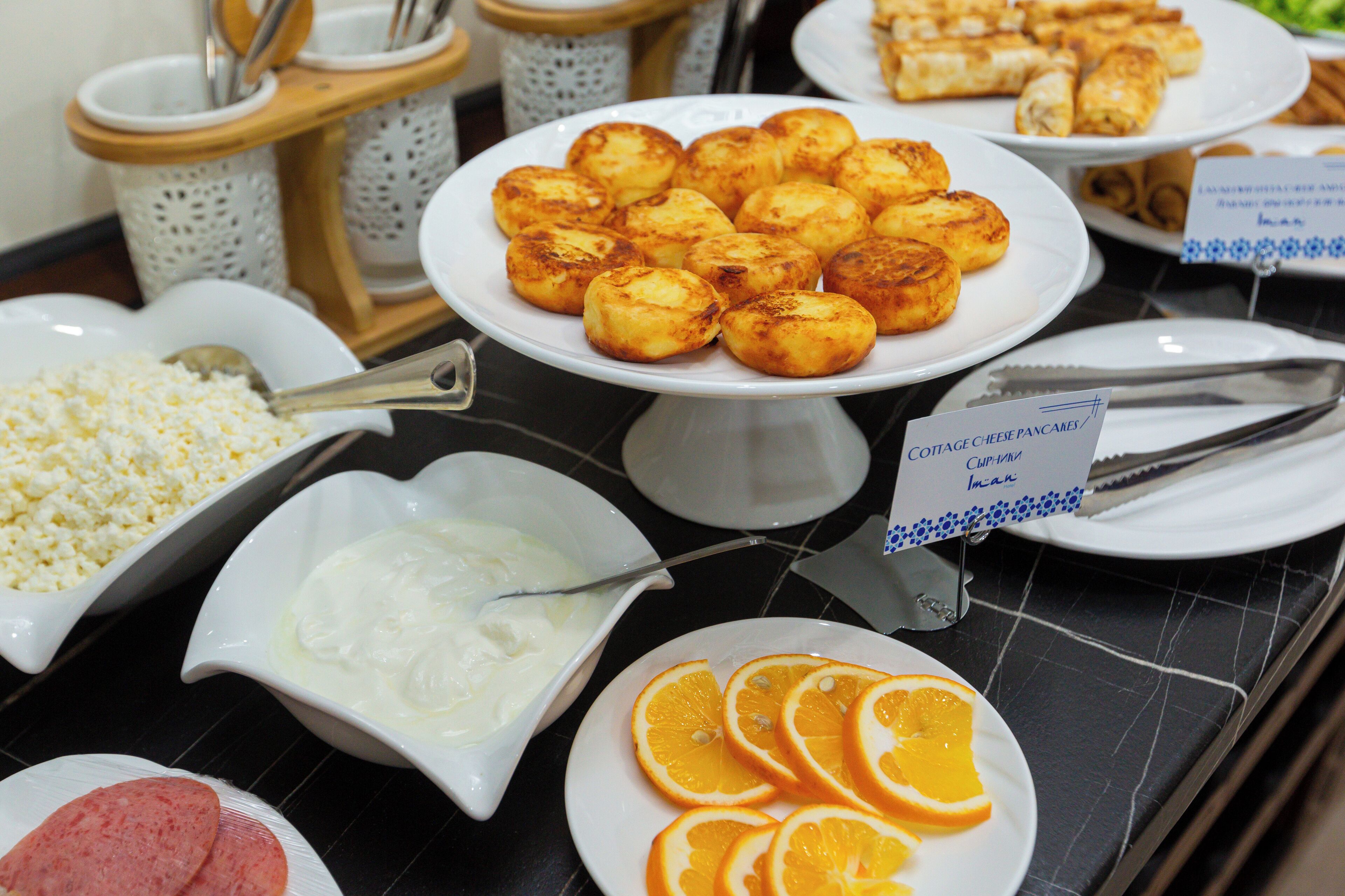 A buffet table with cheese pancakes, yogurt, orange slices, and other food items.