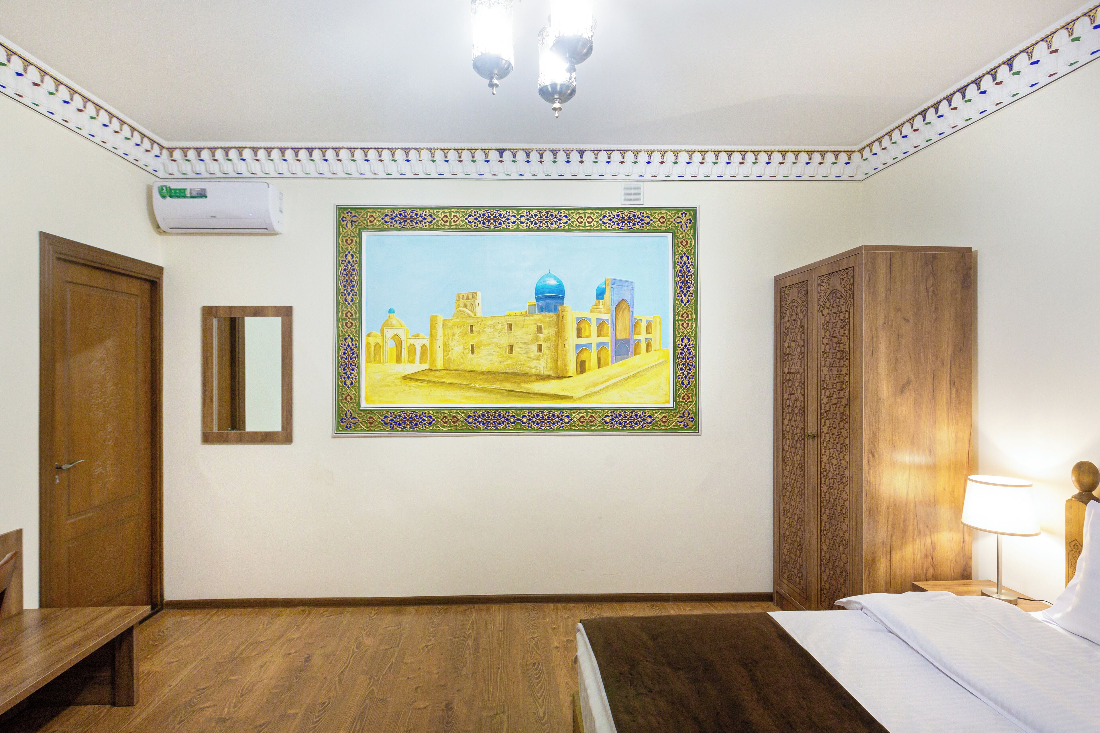 A hotel room with a bed, a wooden bench, a wall-mounted air conditioner, a framed painting of a building, and a wooden wardrobe.