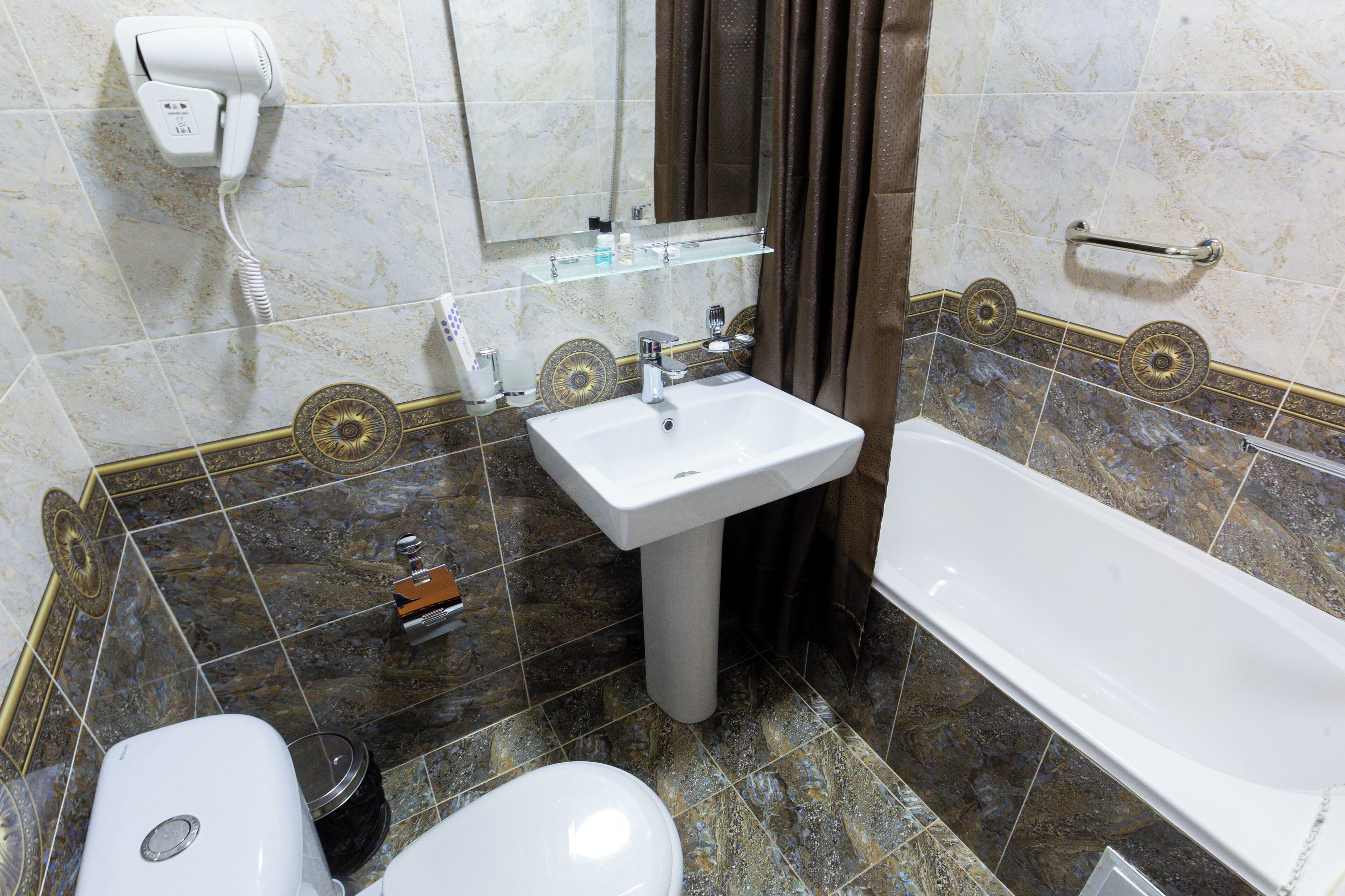 Une salle de bain avec un lavabo blanc, des toilettes et une baignoire. Un sèche-cheveux est fixé au mur et une étagère se trouve au-dessus du lavabo.
