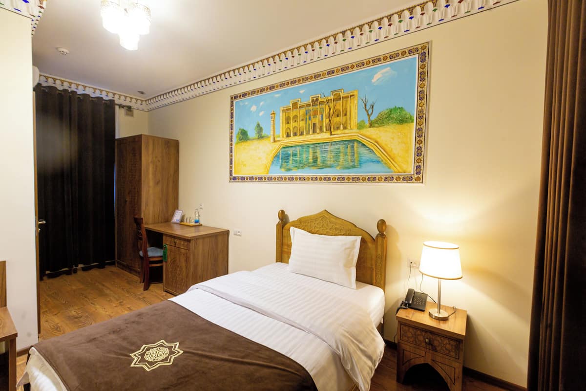 Habitación de hotel con cama, escritorio, silla, mesita de noche con lámpara y un cuadro de un edificio en la pared.