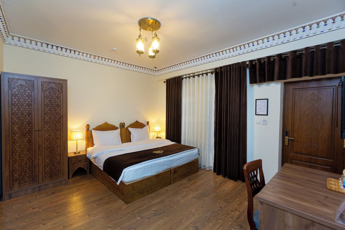 Habitación de hotel con una cama grande, cabecera de madera, dos lámparas de noche, un armario de madera, una mesa de comedor con sillas y un candelabro.