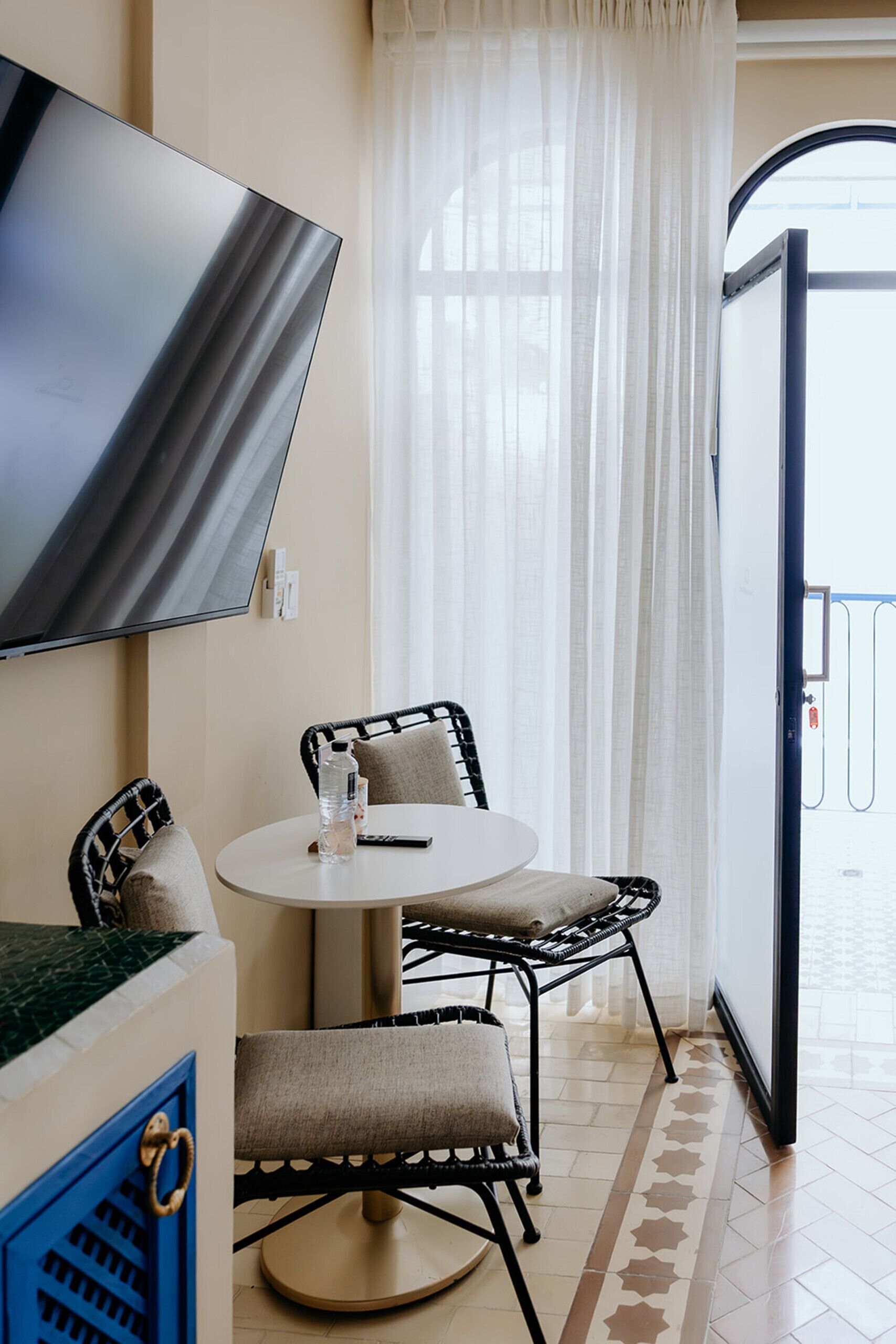 A hotel room with a flat-screen TV mounted on the wall, a round table with two chairs, a small table with a bottle and remote, and a view through a door to the outside.