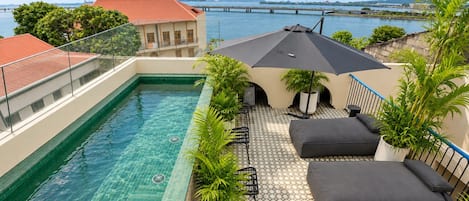 Un área de piscina en la azotea con agua azul cristalina, vista a un puente y un edificio con techo rojo al fondo.
