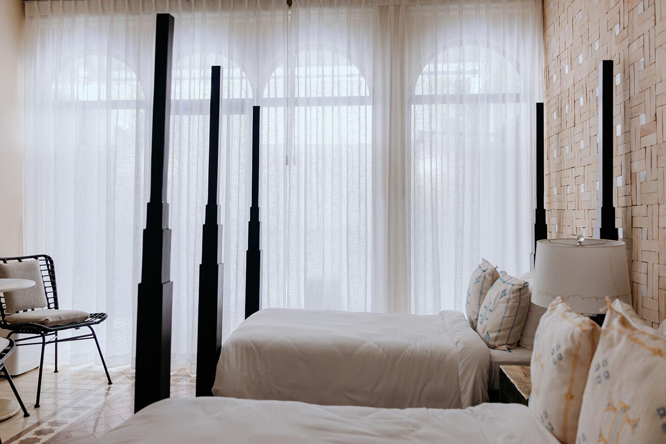 A bedroom with two beds, sheer curtains, a chair, a lamp, and a woven wall.