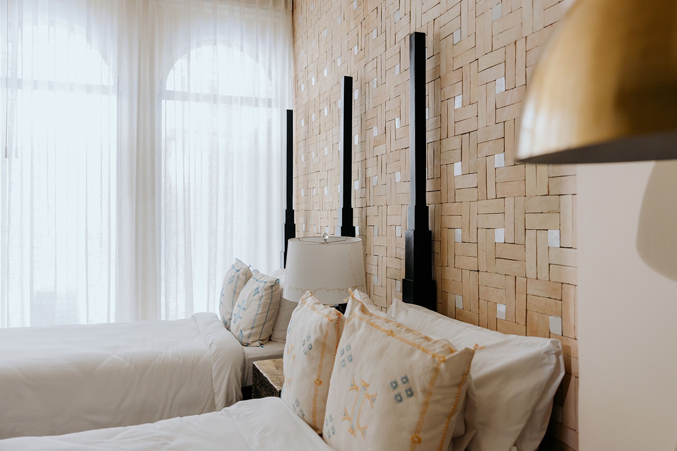 Een hotelkamer met twee bedden, een geweven hoofdbord, een lamp en dunne gordijnen.