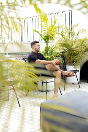 Une personne assise sur une chaise, dans une pièce dont le sol est recouvert de carreaux, avec une plante en pot et une clôture en métal.