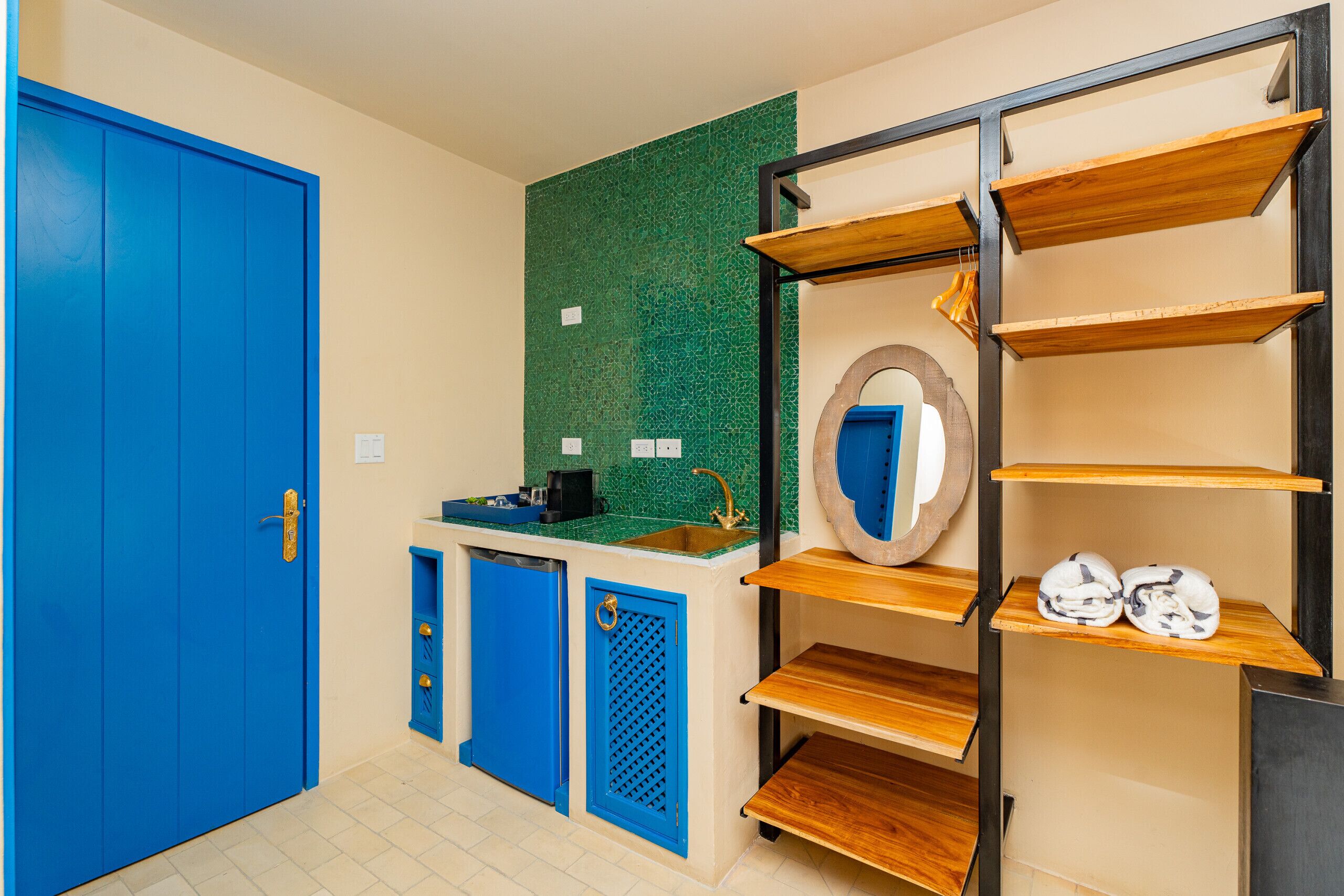 Une pièce avec une porte bleue, un mur recouvert de carreaux verts derrière l’évier, un évier, un miroir et des étagères en bois.