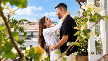 Un couple s’embrasse sur un balcon, avec vue sur des bâtiments et de la verdure.