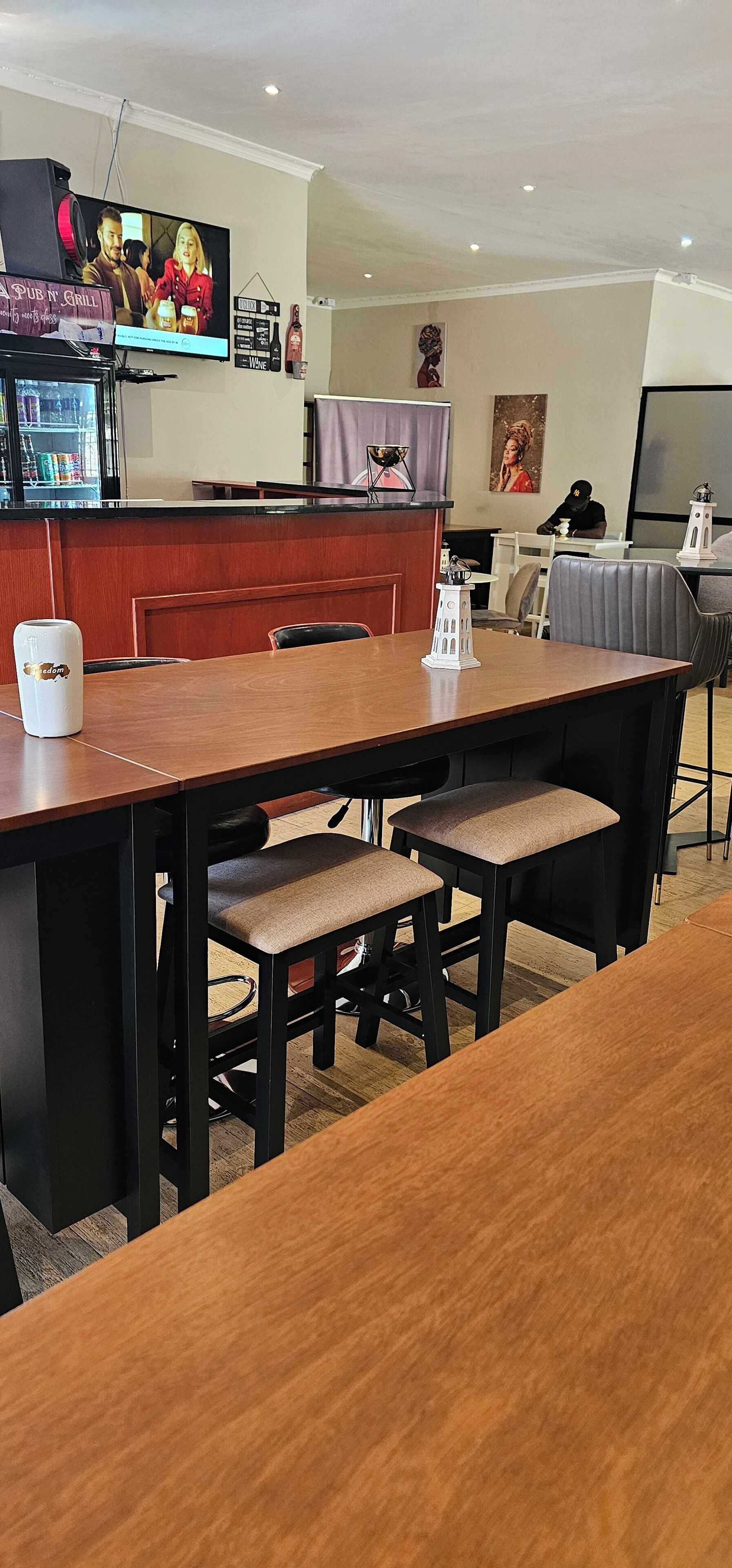 A dining area with a long wooden table, cushioned chairs, a small white table lamp, and a television mounted on the wall.