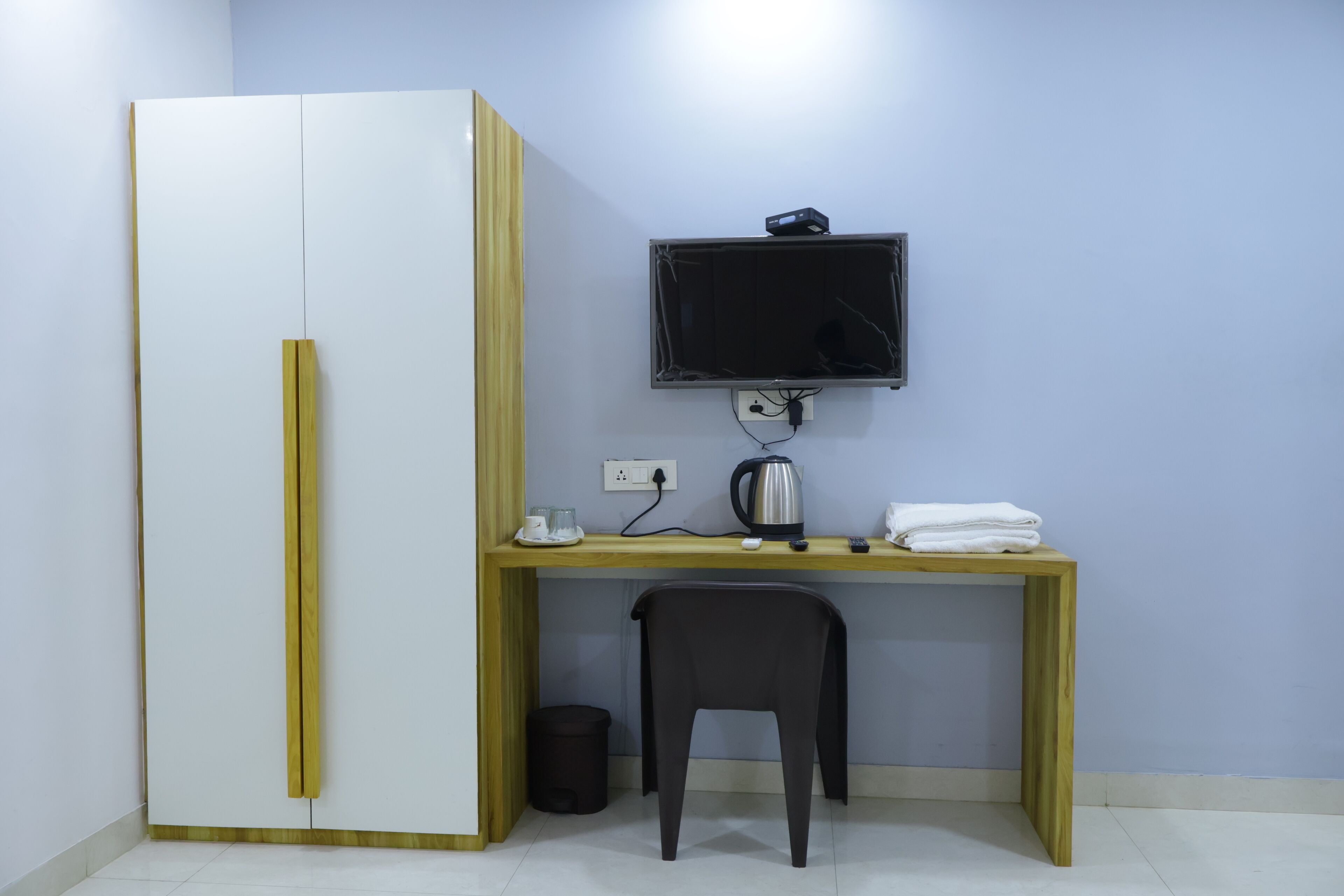 A room with a wooden desk, a television mounted on the wall, a wardrobe, and a trash bin.