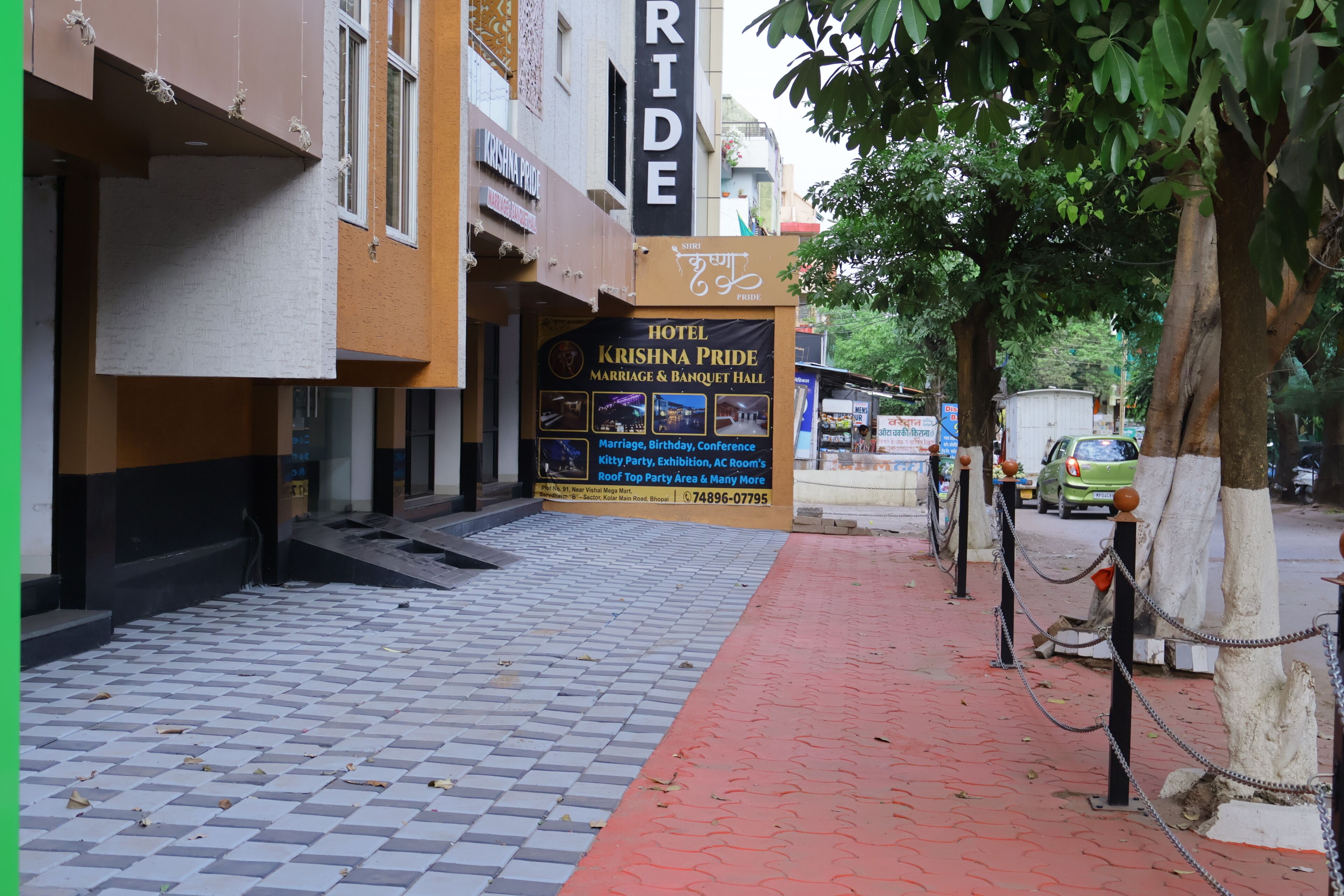 Entrada de un hotel con un camino de baldosas, un cartel de Krishna Pride Marriage & Banquet Hall, y un vehículo estacionado.