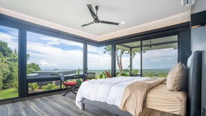 Een slaapkamer met een groot raam, een bed, een plafondventilator en uitzicht op de omgeving.