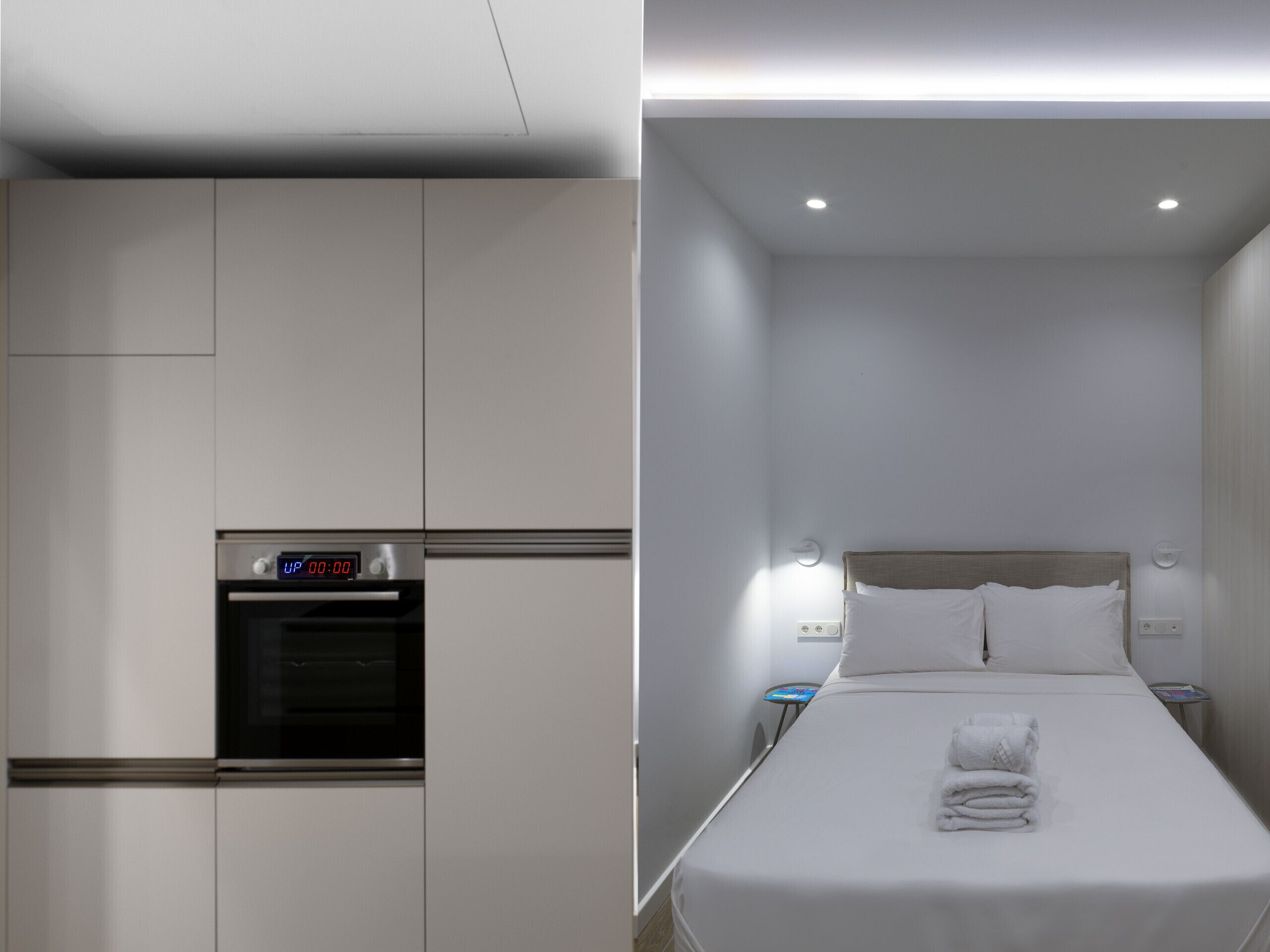 A modern hotel room with a kitchenette, a bed with white linens, and a bedside table.