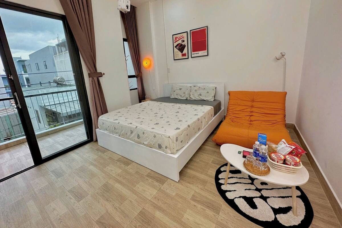 Ein modernes Schlafzimmer mit Bett, Sofa, einem kleinen Tisch mit Gegenständen und Blick auf Gebäude durch ein Fenster.