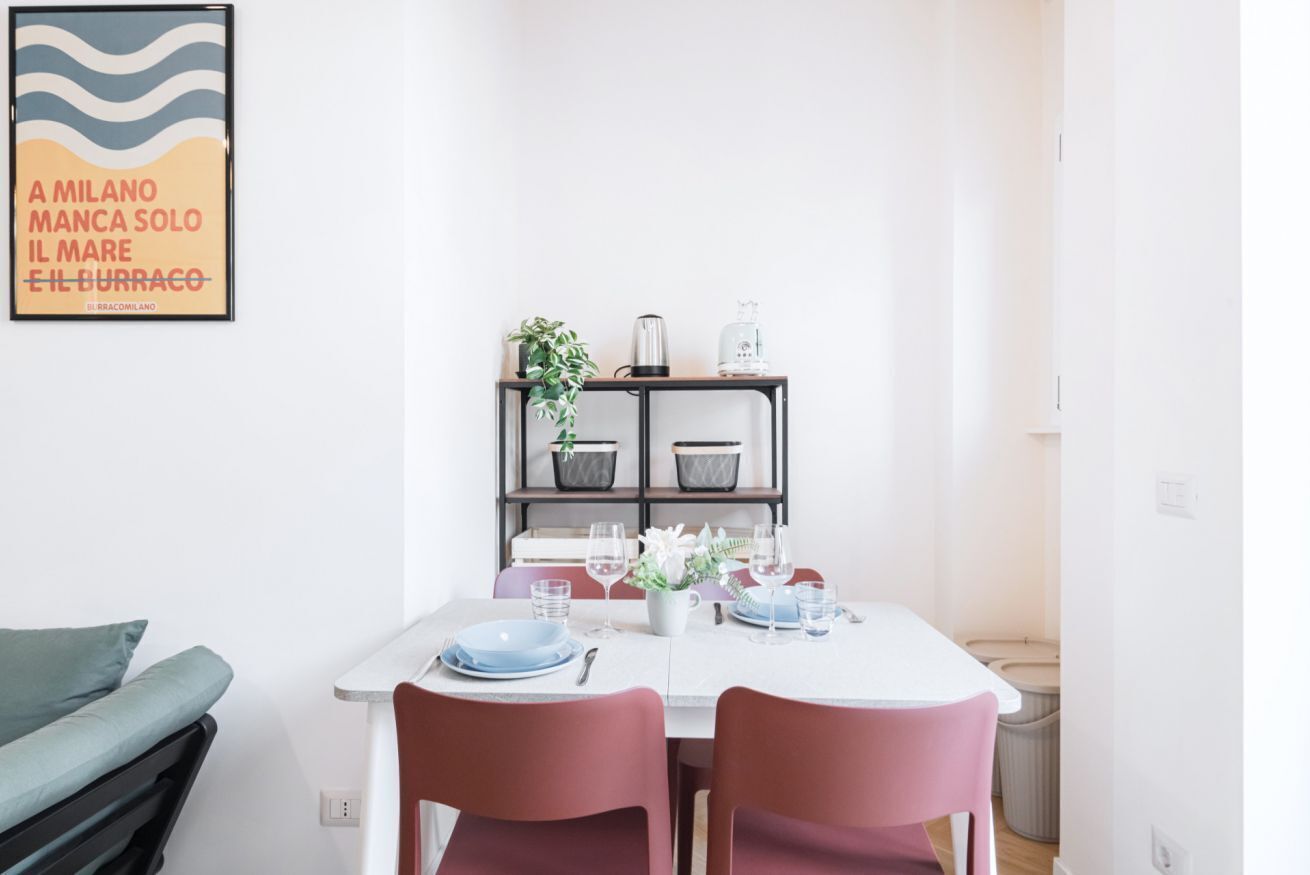 A dining area with a white table set for two, red chairs, a potted plant, and a coffee machine.