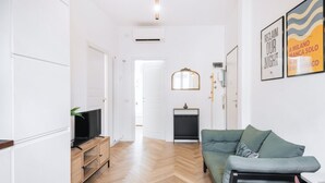 A modern living room with a wooden herringbone floor, a green sofa, a TV on a wooden stand, and a mirror on the wall. - Easylife - Apartment with balcony near Navigli (Milano)