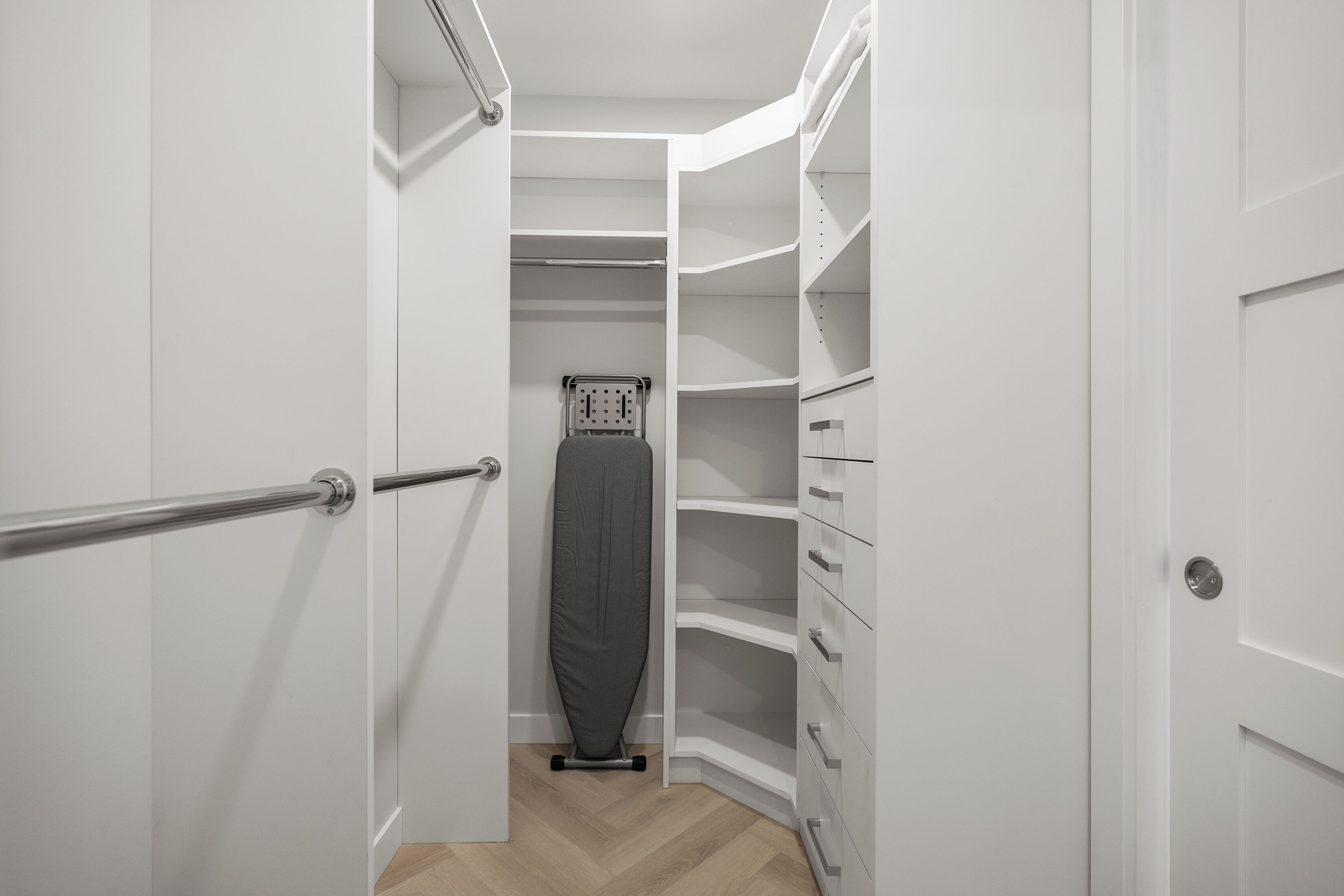 Um closet pequeno e estreito, com paredes brancas, um centro de passar roupa de metal e um sapateiro.
