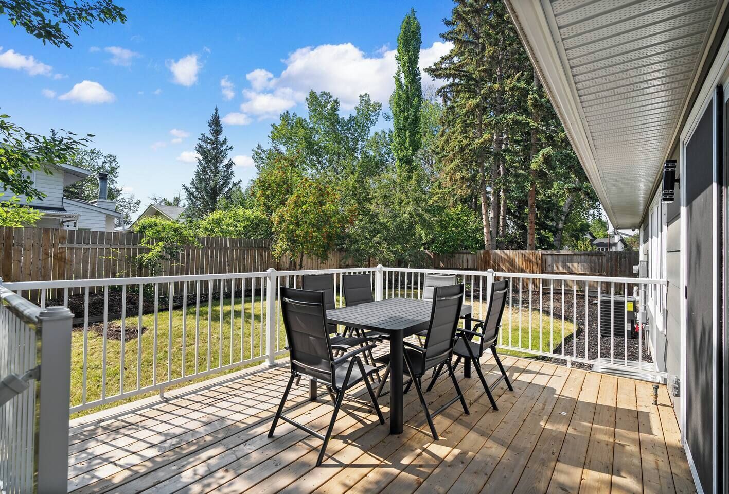 Une terrasse avec une table et des chaises, entourée d’une clôture blanche, donnant sur un jardin avec des arbres et une clôture en bois.