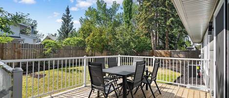 A deck with a table and chairs, surrounded by a white fence, overlooking a backyard with trees and a wooden fence.