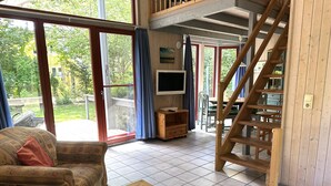 A spacious living area with a sofa, a glass-top coffee table, a TV, and a wooden staircase. - Wildgans 83 - Classic Scandinavian vacation home for up to 6 people (Mirow)