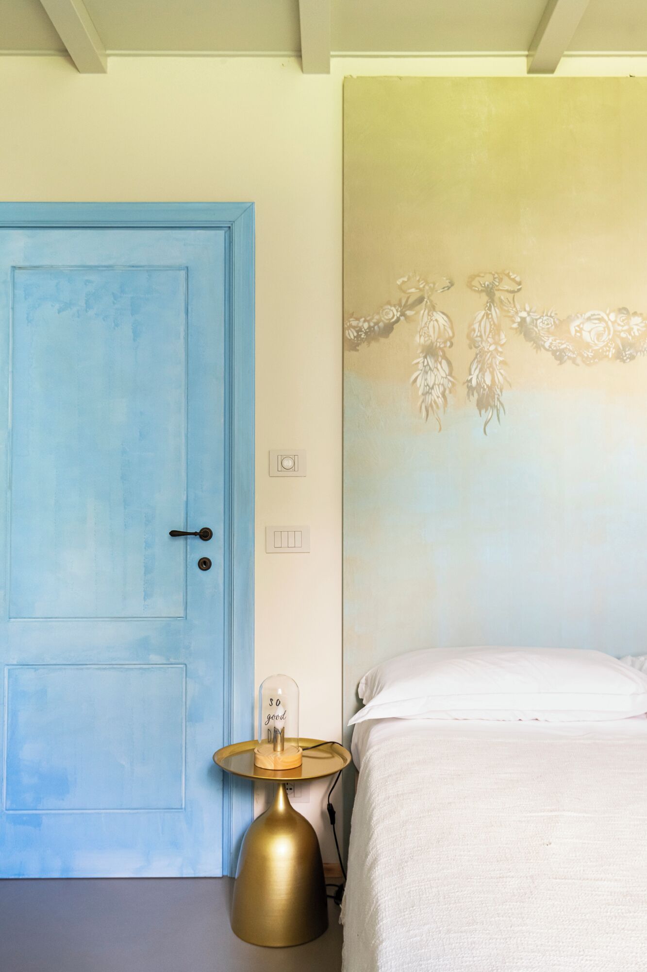 A room with a blue door, a bed with white bedding, a bedside table with a lamp, and a wall with floral wallpaper.