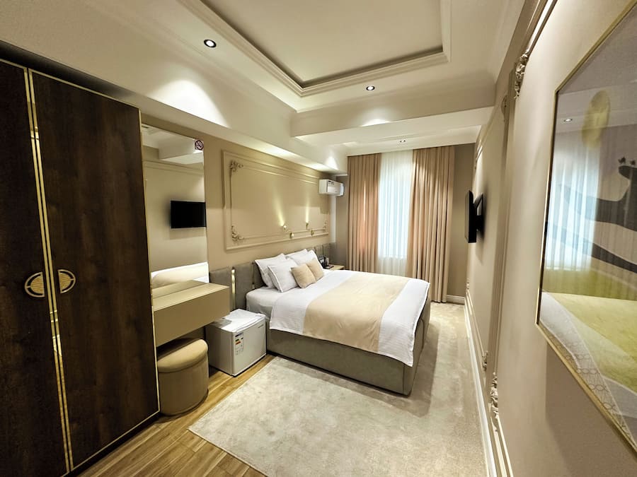 A modern hotel room with a large bed, a vanity with a mirror, a small seating area, and a large window with curtains.