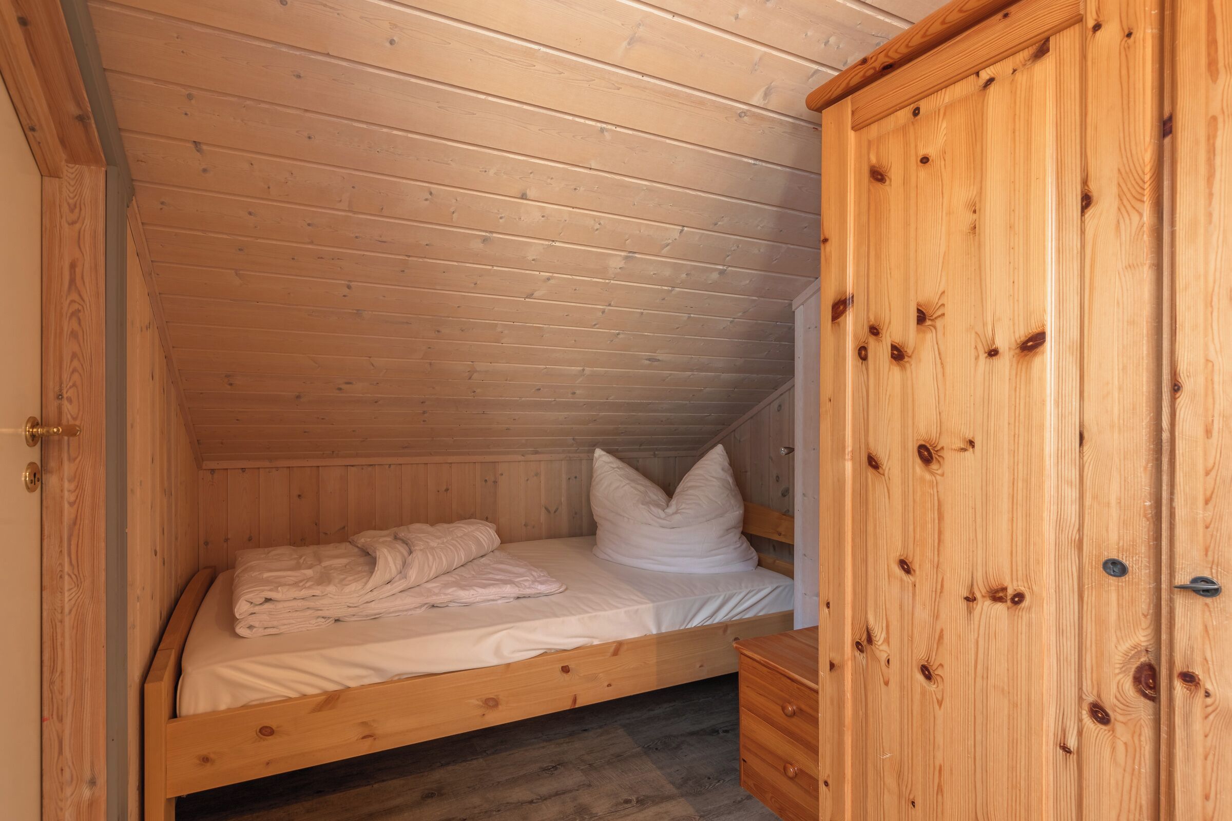 Una habitación pequeña de madera con una cama individual, un armario de madera y una puerta.