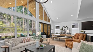 A spacious living area with a large window, a sofa, a coffee table, and a dining area with a bench and table. - Harmony Haven - EV - Hot Tub - Pups Allowed (Cle Elum)