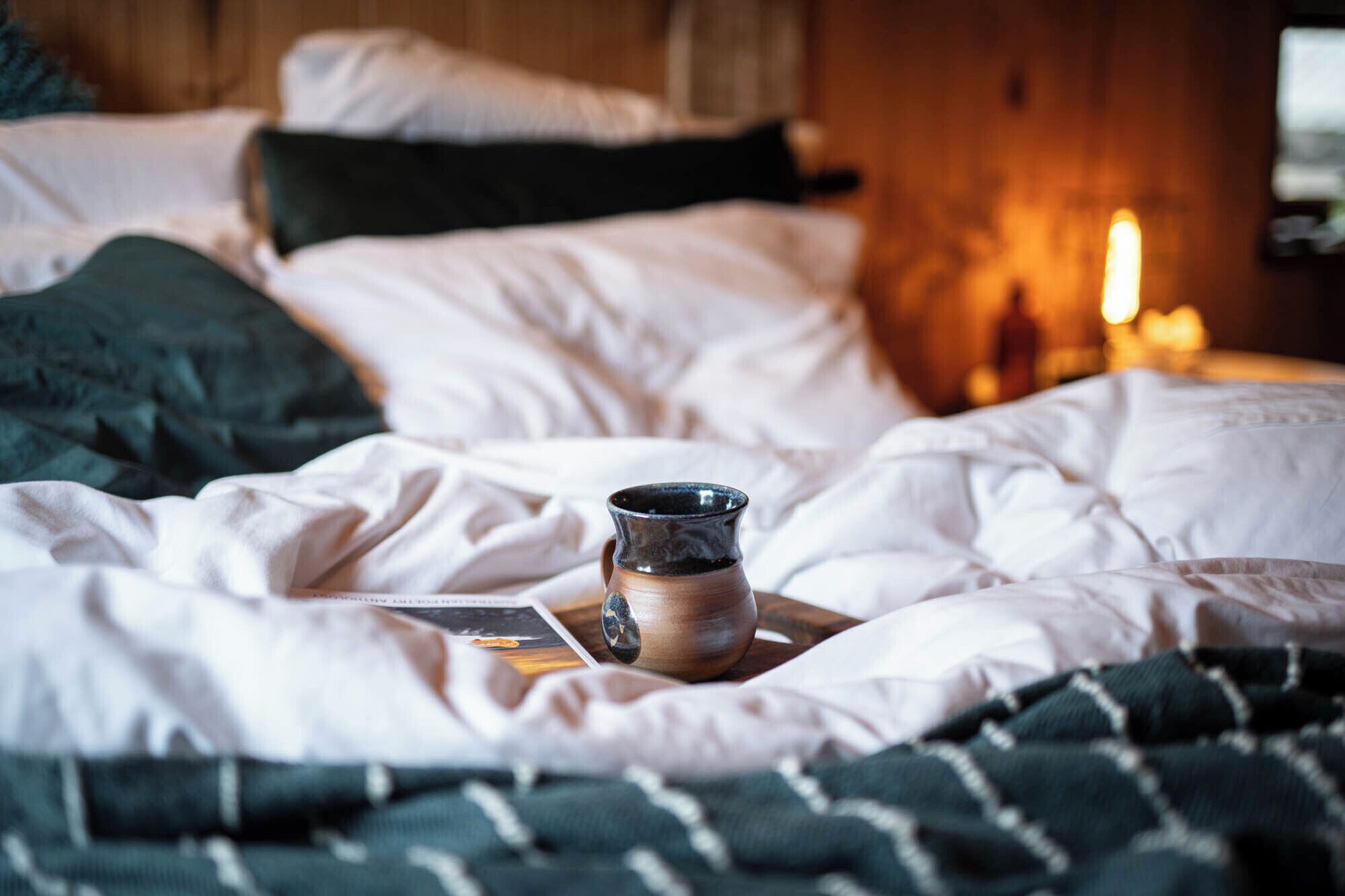 A bed with white bedding and a small ceramic mug on a wooden tray.