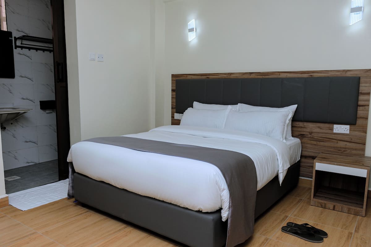 A modern hotel room with a large bed, a wooden headboard, a small bedside table, and a shower area.