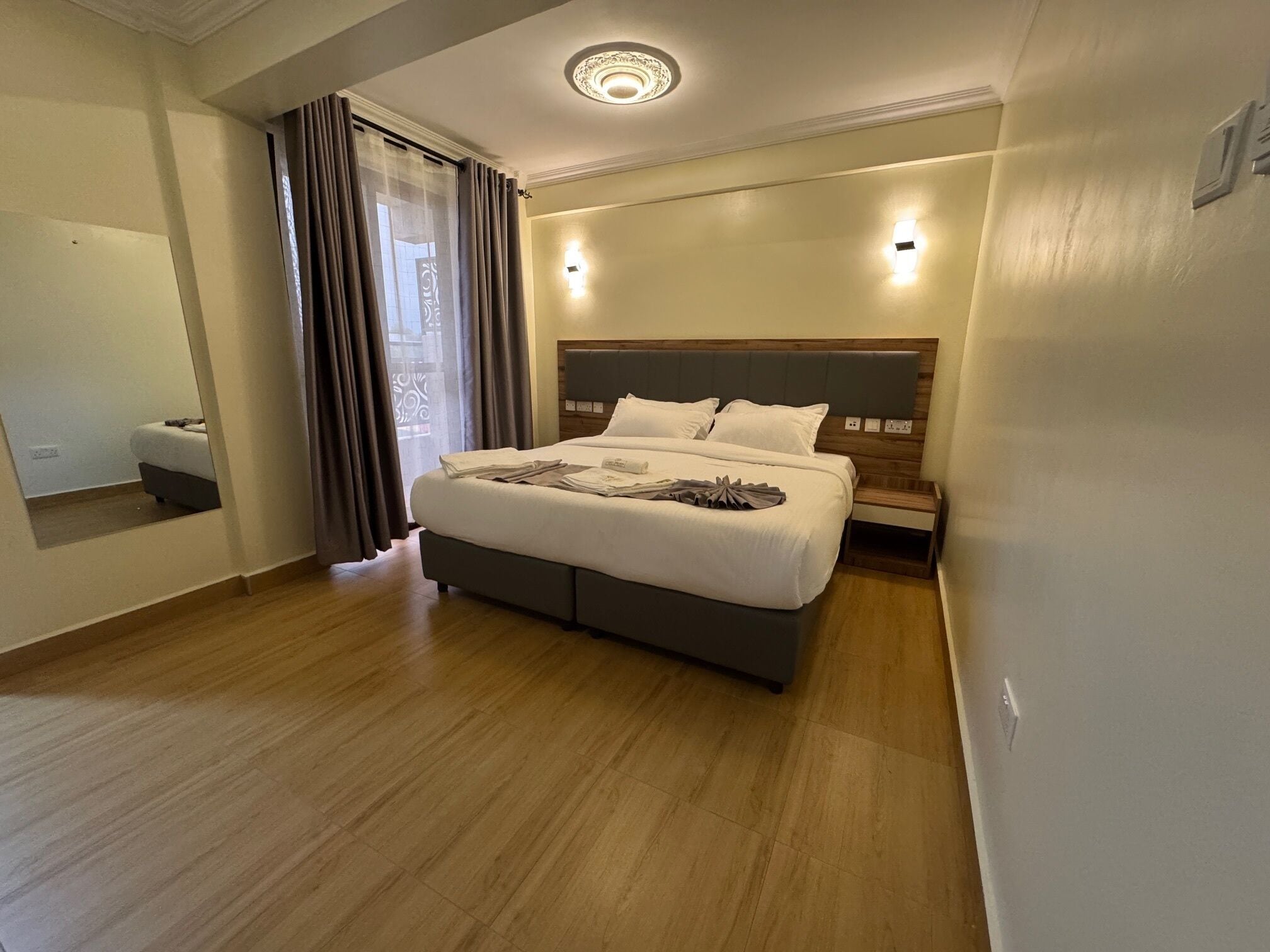Una habitación de hotel moderna con una cama grande, suelo de madera, cabecero con iluminación integrada y un espejo en una pared.