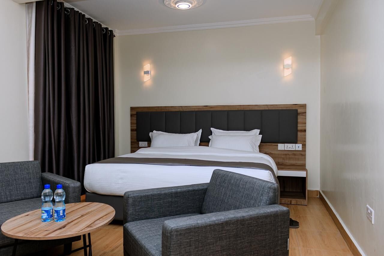 Une chambre d’hôtel avec un grand lit, des fauteuils gris rembourrés, une petite table en bois avec des bouteilles d’eau et deux lampes fixées au mur.